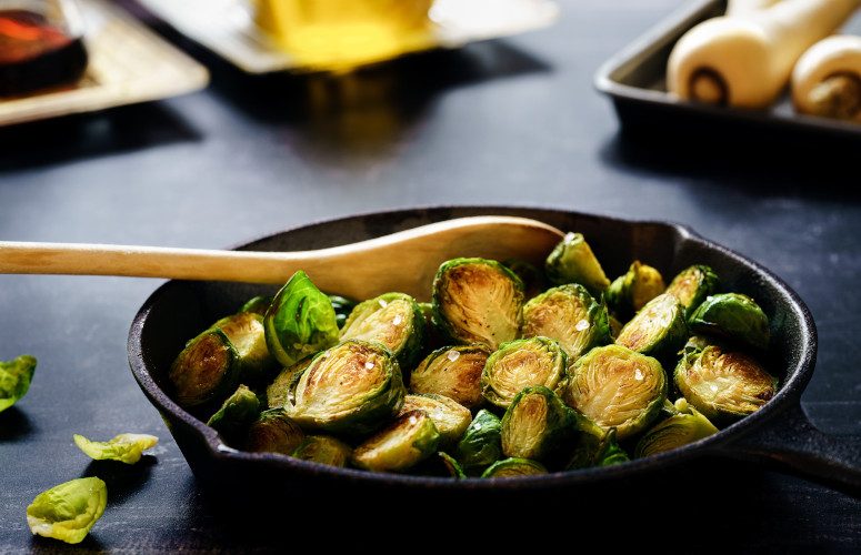 Cast iron pan with fried brussel sprouts and a wooden spoon resting in it.