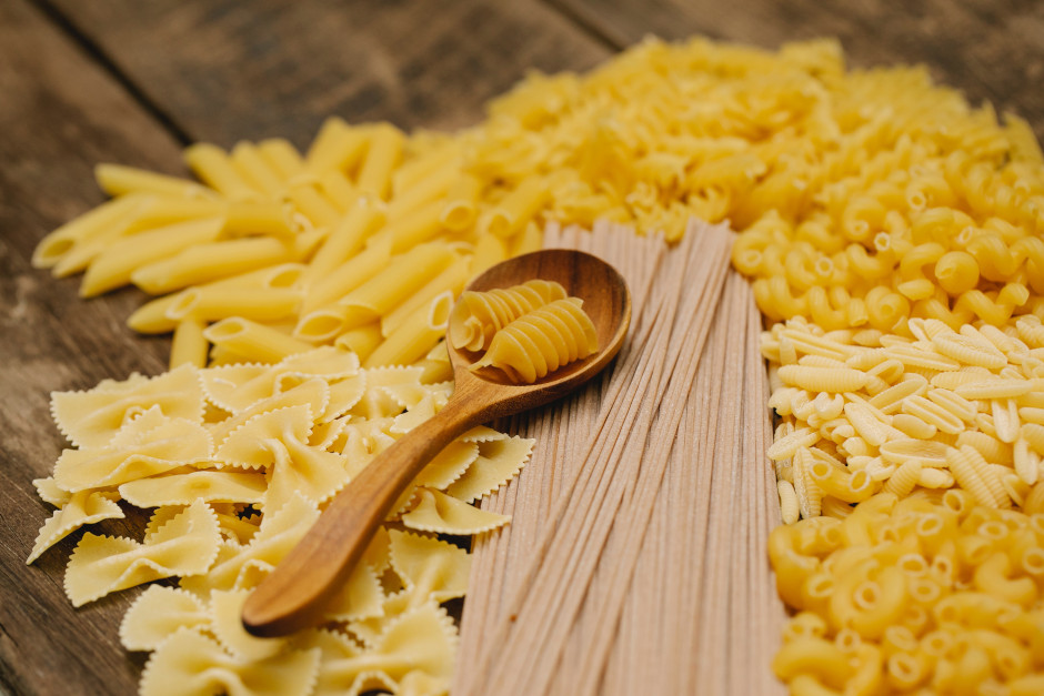 Different types of pasta on a wood counter with a wooden spoon on top.